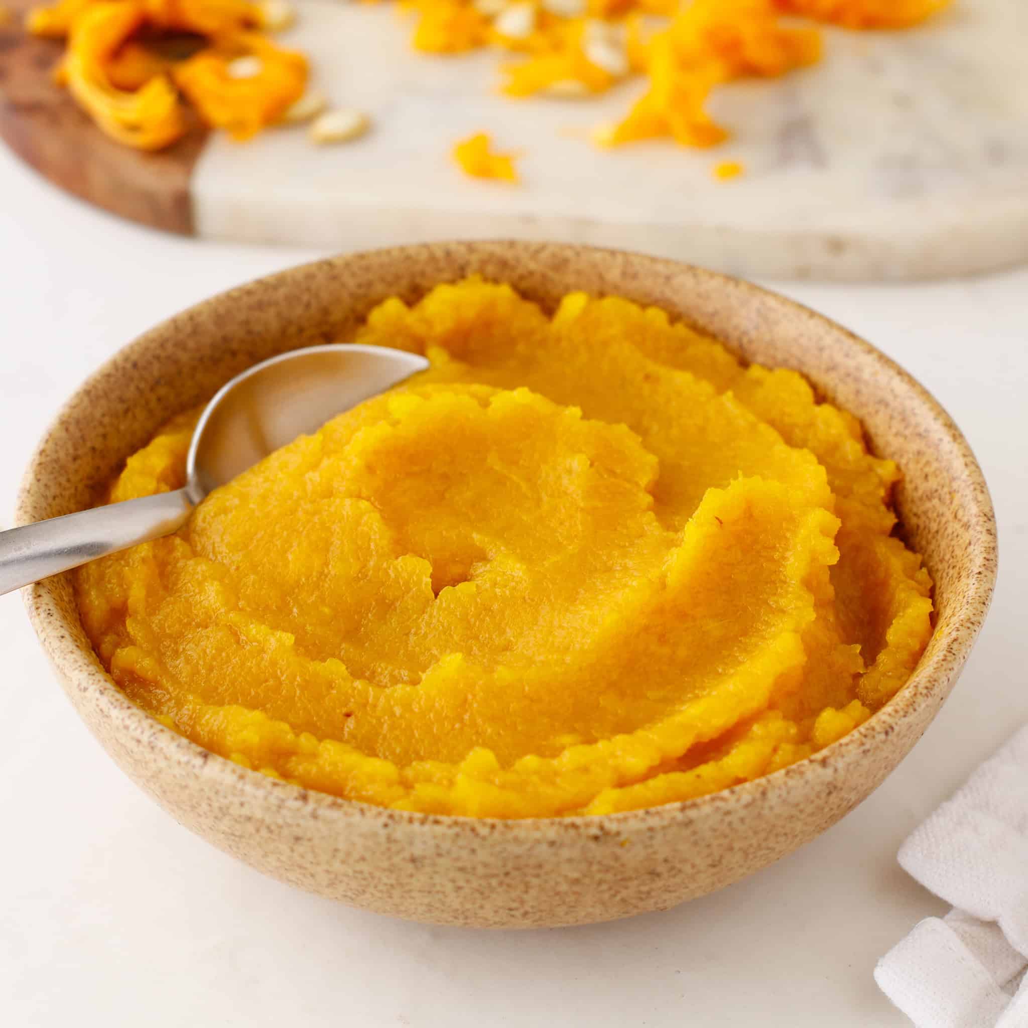 A bowl of smooth pumpkin puree with a spoon, on a white surface near sliced pumpkin pieces. This creamy pumpkin puree is perfect for seasonal recipes or enjoying on its own.