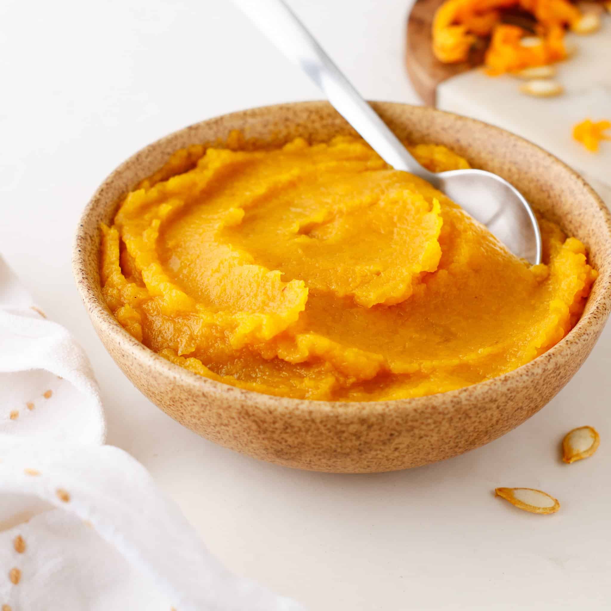 A bowl of smooth orange pumpkin puree with a spoon, on a white surface next to a napkin and sliced vegetables, makes a simple and delicious way to enjoy the rich flavor of Pumpkin Puree.
