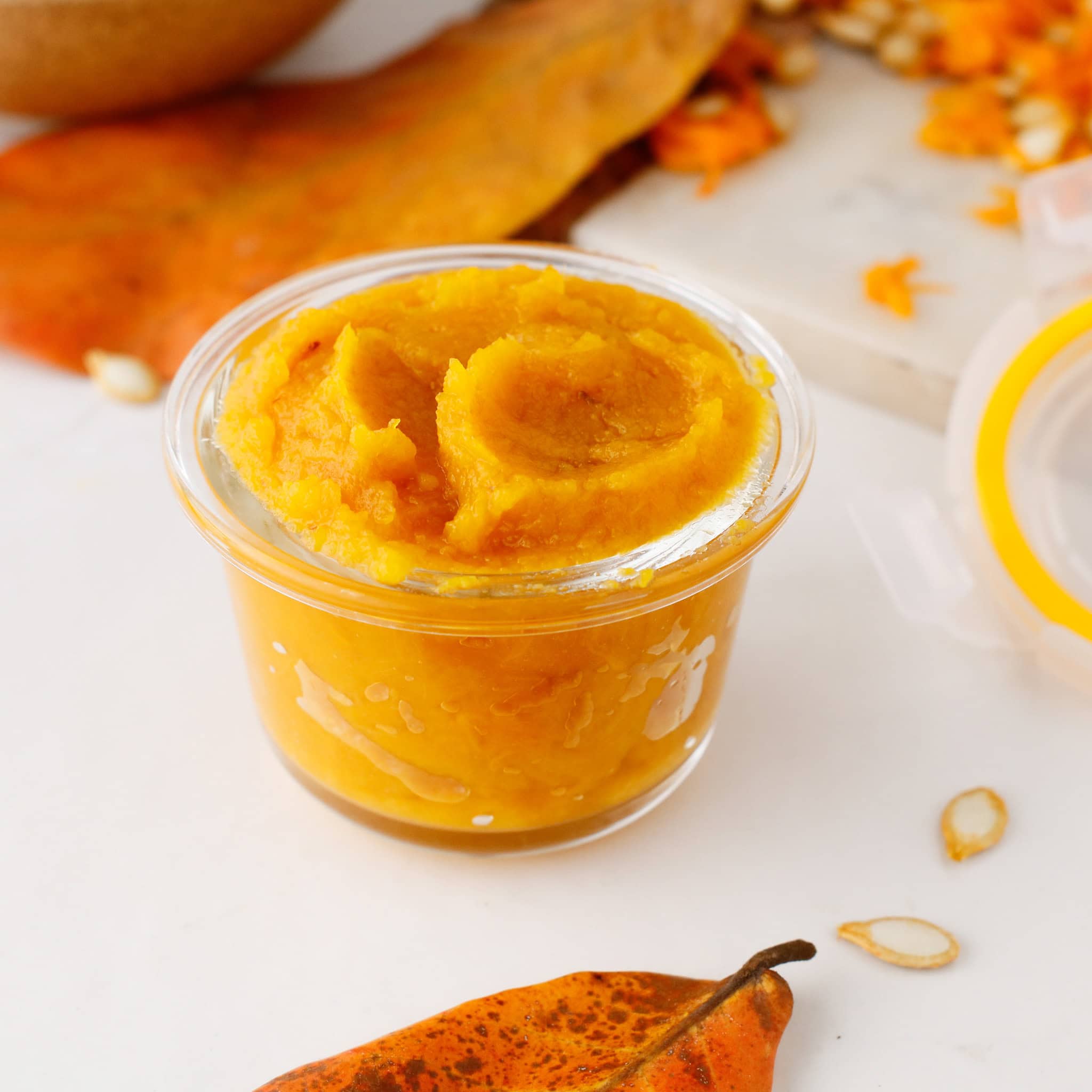 A small clear container filled with vibrant pumpkin puree, surrounded by autumn leaves and pumpkin seeds.