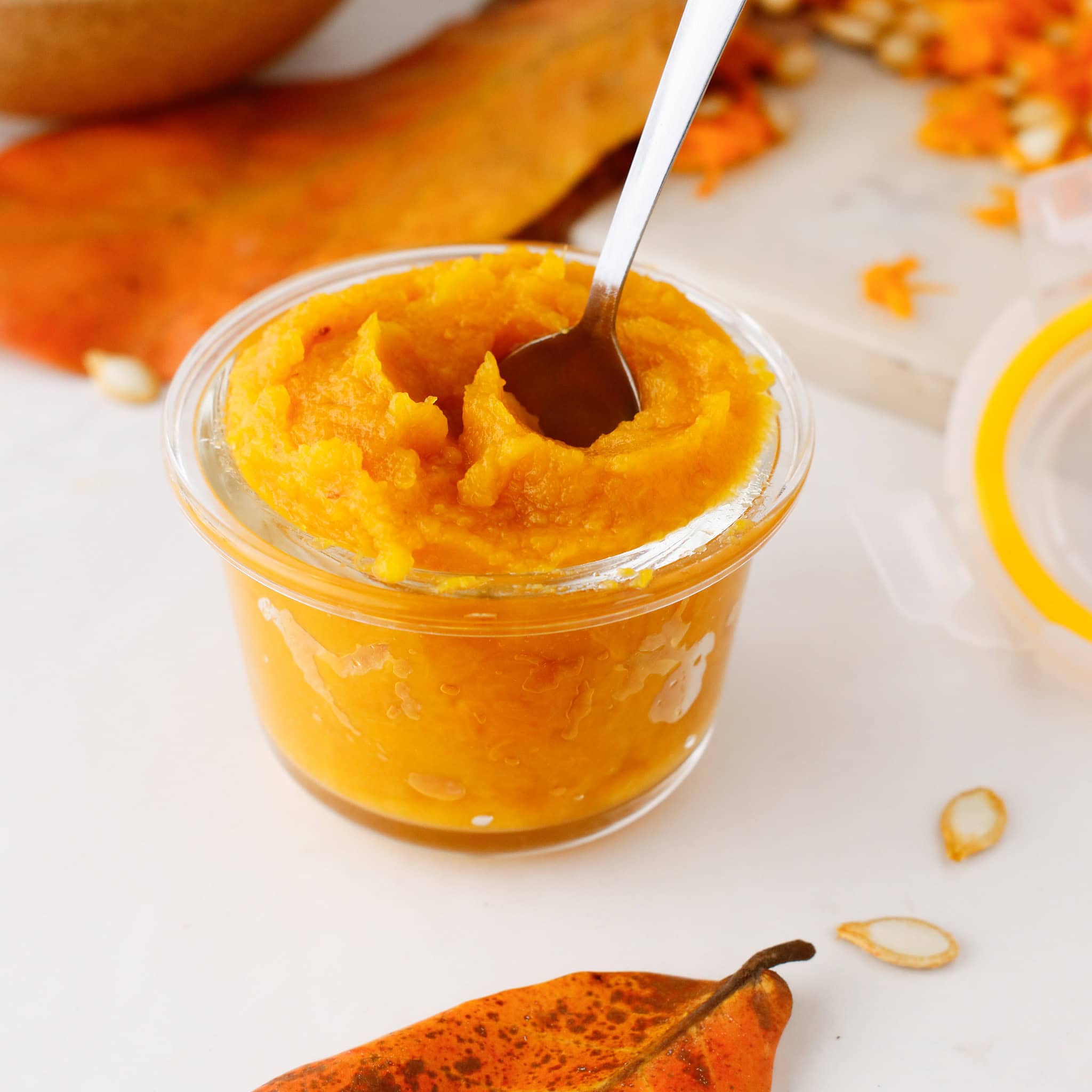 A glass jar of Pumpkin Puree with a spoon, surrounded by autumn leaves and pumpkin seeds, captures the cozy flavors of fall.