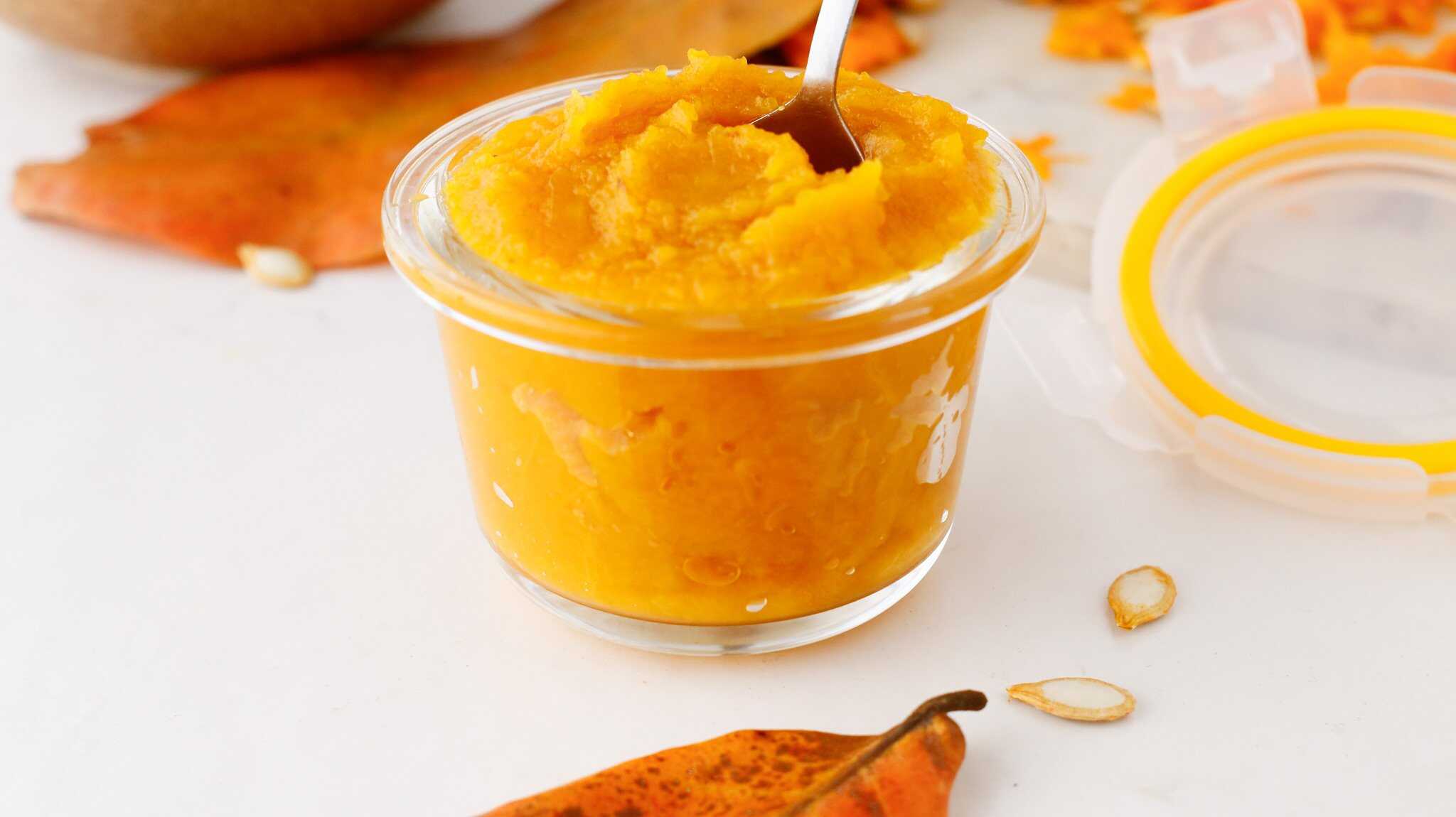 A small glass jar filled with rich pumpkin puree, a spoon inside, surrounded by autumn leaves and seeds.