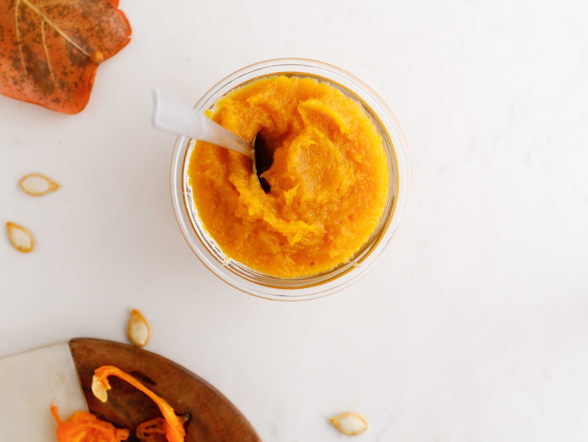 A glass jar filled with smooth pumpkin puree, with a spoon inside, sits on a white surface.