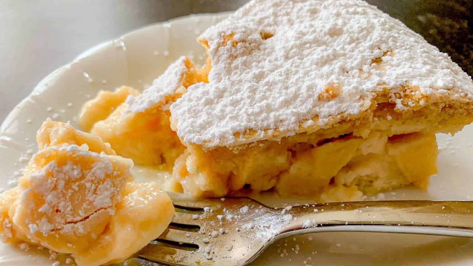 A slice of creamy pie topped with powdered sugar sits on a white plate. A fork with a small piece of the pie rests beside it, and the plate is sprinkled with sugar.