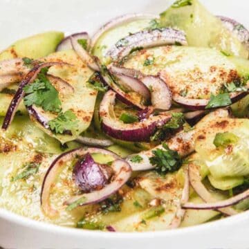 Sliced cucumber salad with red onions, cilantro, and spices in a white bowl with a fork on the side.