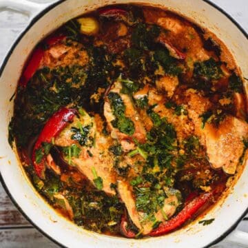 A pot of fish stew with herbs, red peppers, and green leafy vegetables in a white enamel pot.