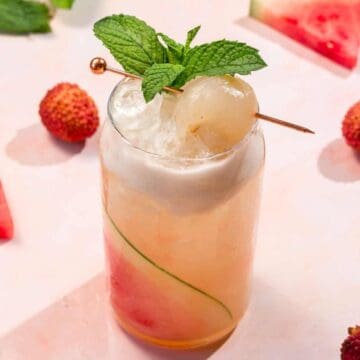 A tall glass of lychee and watermelon cocktail with ice, mint garnish, and lychees, surrounded by fruit slices.