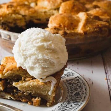 A slice of apple pie with a scoop of vanilla ice cream on a plate, with the whole pie in the background.