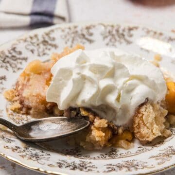 A serving of peach cobbler topped with whipped cream on a decorative plate with a spoon.