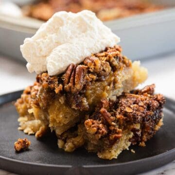 A slice of pecan pie topped with whipped cream on a black plate, with a baking dish in the background.