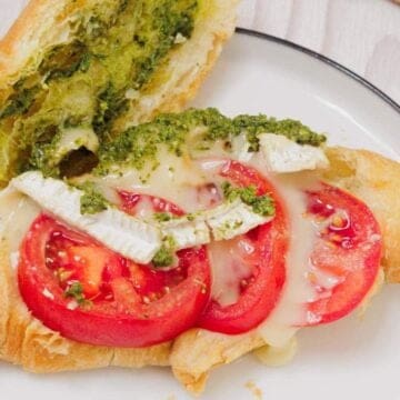 A croissant sandwich with tomato slices, brie cheese, and pesto on a white plate.
