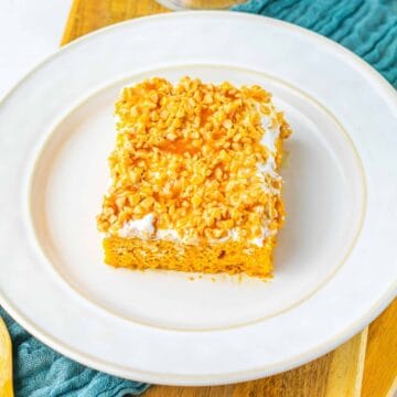 A square slice of nut-topped cake with icing on a white plate, next to a wooden spoon and blue napkin.