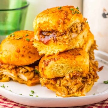 Three BBQ chicken sliders stacked on a white plate, with a red checked cloth and a green glass nearby.
