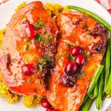 Chicken breasts with cranberry sauce, green beans, and stuffing on a white plate on a red-checkered tablecloth.