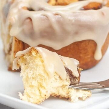 A cinnamon roll with icing, with a bite taken out on a fork, on a white plate.