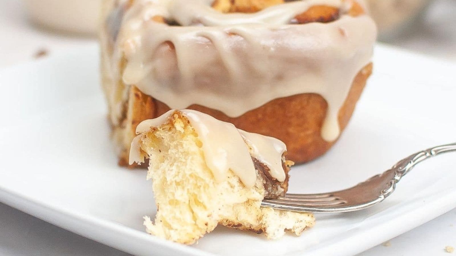 A cinnamon roll with icing, with a bite taken out on a fork, on a white plate.