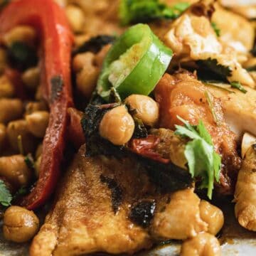 Close-up of a plate with cooked chicken, chickpeas, red peppers, herbs, and sliced jalapeños.