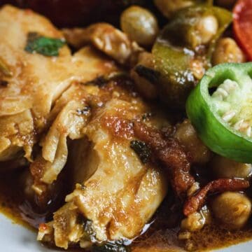 Close-up of cooked chicken with chickpeas, sliced green chili, tomato, and sauce on a white plate.
