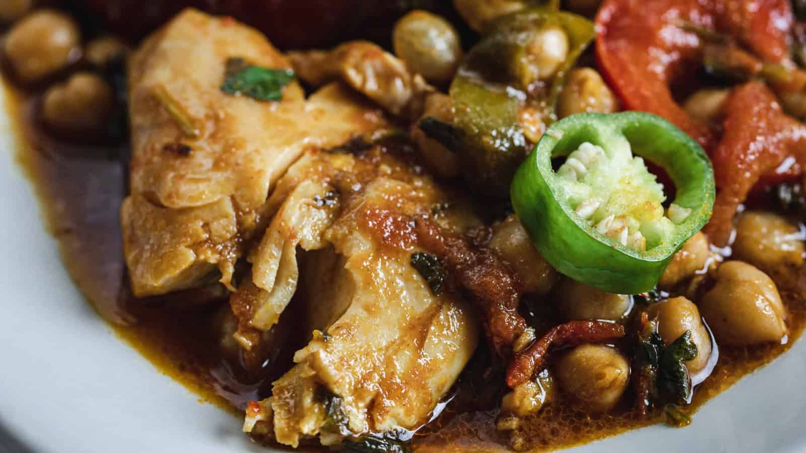 Close-up of cooked chicken with chickpeas, sliced green chili, tomato, and sauce on a white plate.