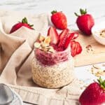 A jar of Strawberry Almond Overnight Chia Oats topped with strawberries and sliced almonds, surrounded by fresh strawberries.