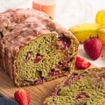 Sliced loaf of strawberry banana bread with strawberries and bananas in the background on a wooden board.