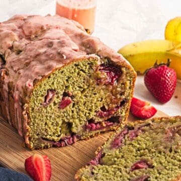 Sliced loaf of strawberry banana bread with strawberries and bananas in the background on a wooden board.