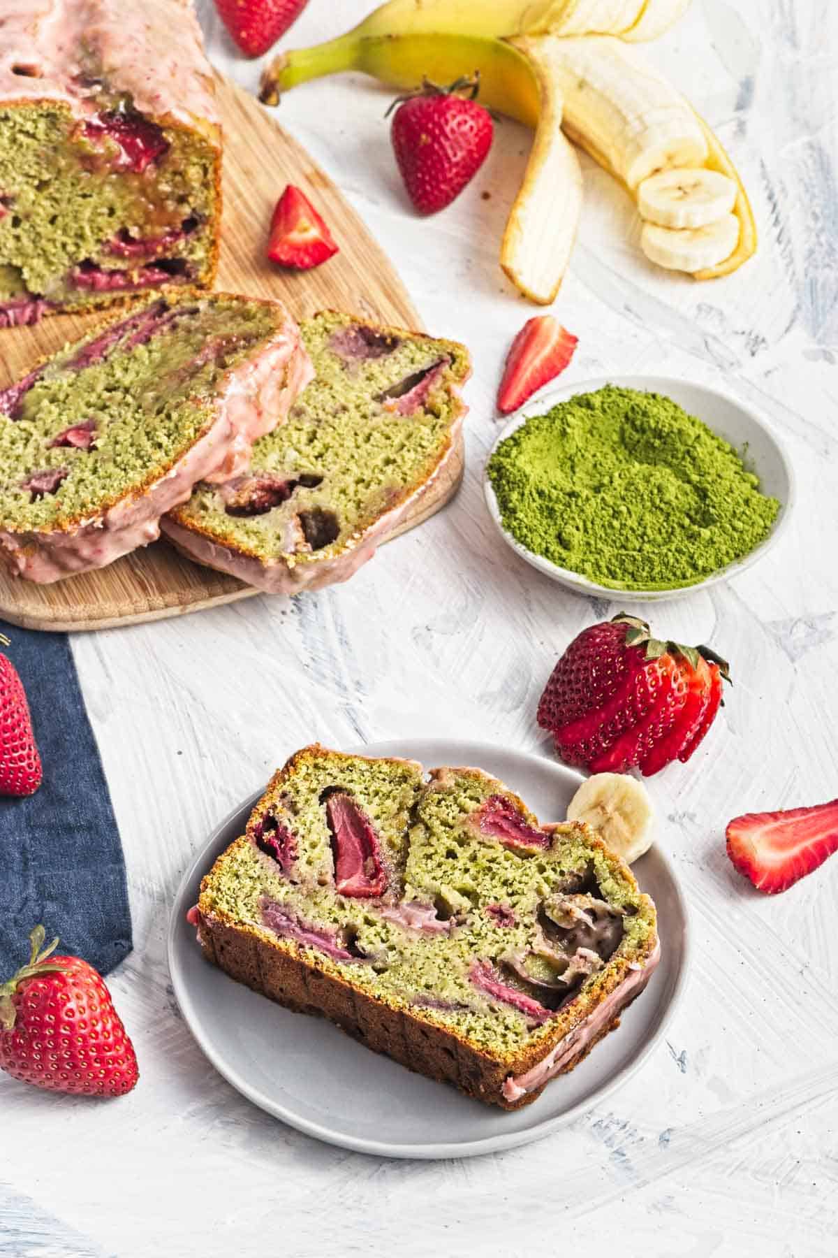 A slice of matcha strawberry banana bread on a plate, with fresh strawberries, banana, and matcha powder nearby.