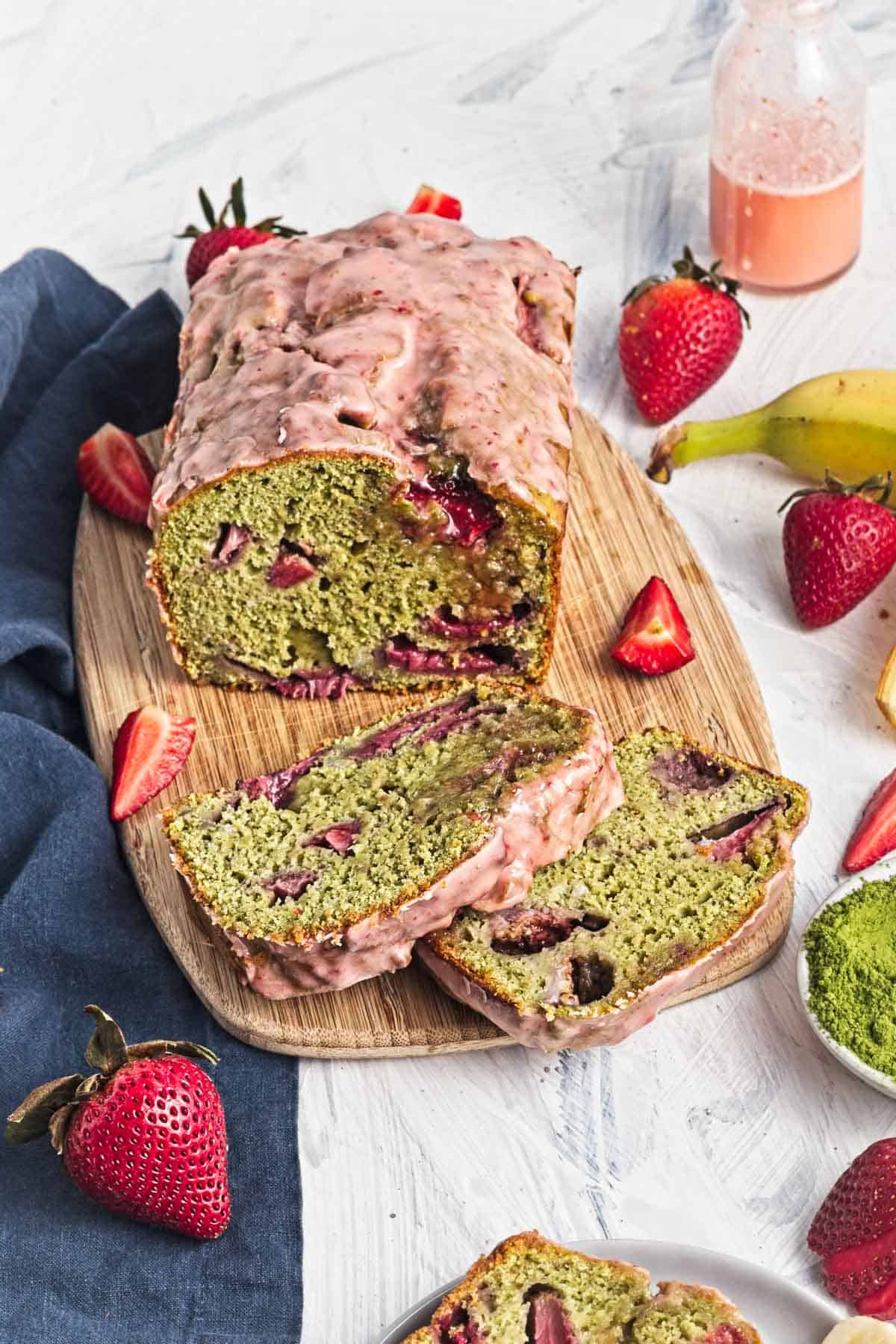 Sliced matcha strawberry loaf with pink glaze on a wooden board, surrounded by fresh strawberries.