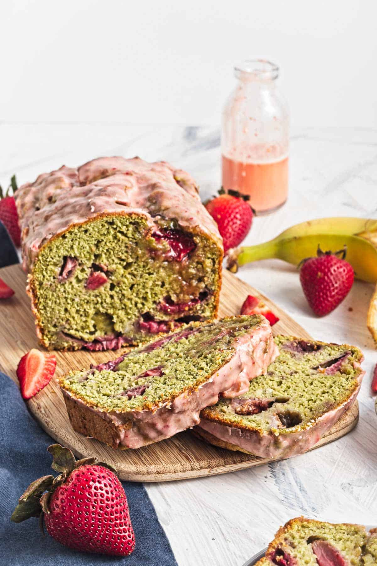 Sliced strawberry banana bread with pink glaze, fresh strawberries, and a banana on a wooden board.
