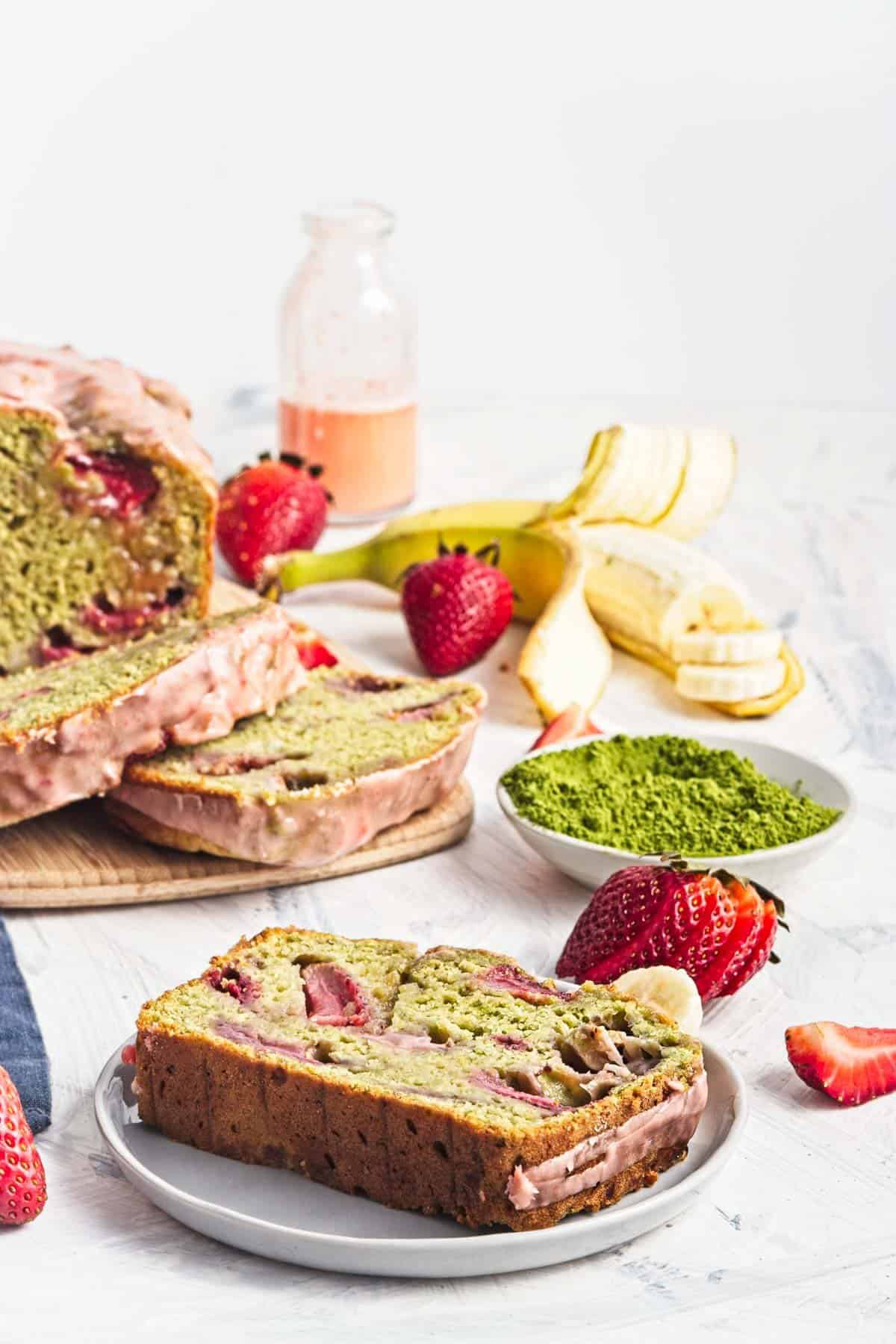 Sliced matcha strawberry banana bread on a plate with fresh strawberries and bananas nearby.