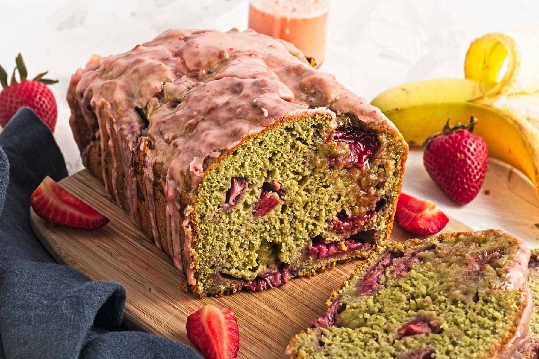 A sliced loaf of banana bread with strawberries, topped with pink glaze, on a wooden board with fruit nearby.