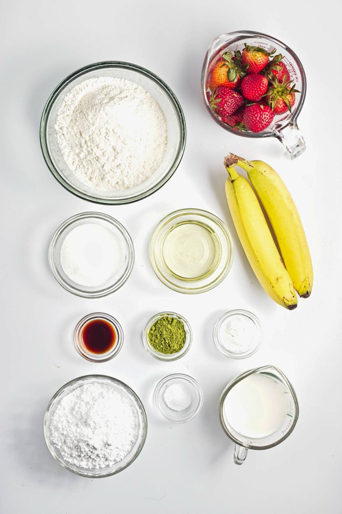 Bowls of flour, sugar, oil, matcha, vanilla, milk, baking powder, salt, strawberries, and two bananas on a white background.
