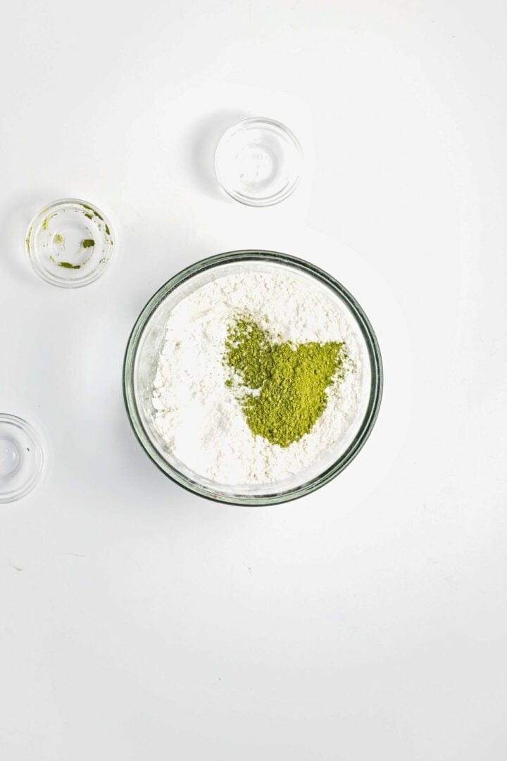 A glass bowl with flour and matcha powder, surrounded by small empty glass bowls on a white surface.