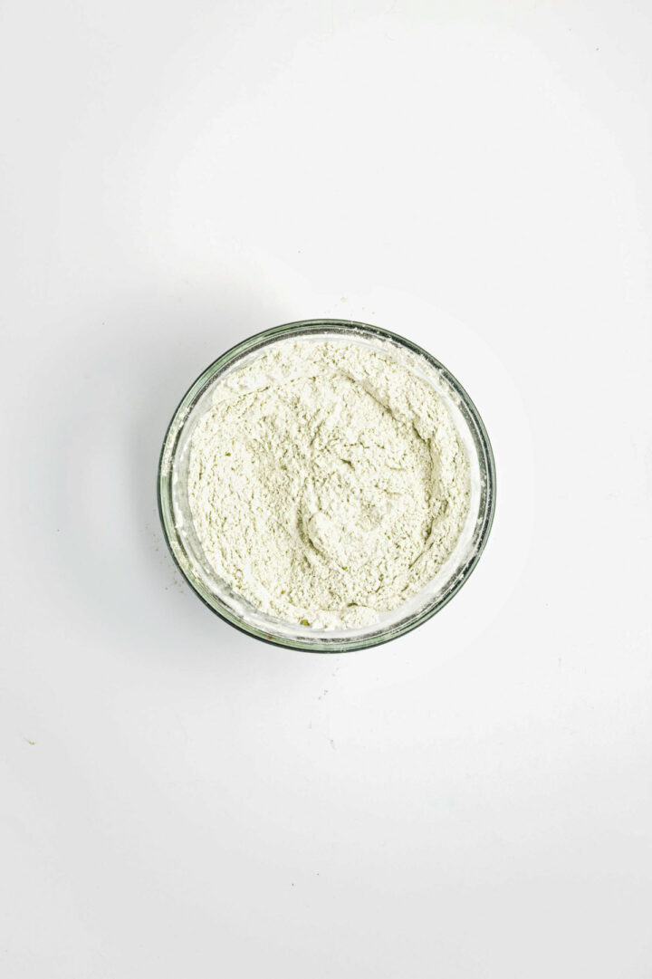 A glass bowl filled with white flour on a plain white background.