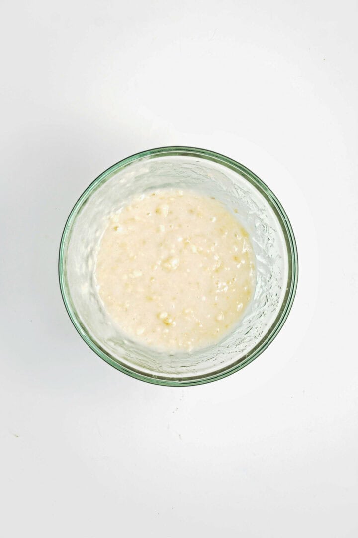 A glass bowl filled with lumpy, pale batter mixture on a white surface.