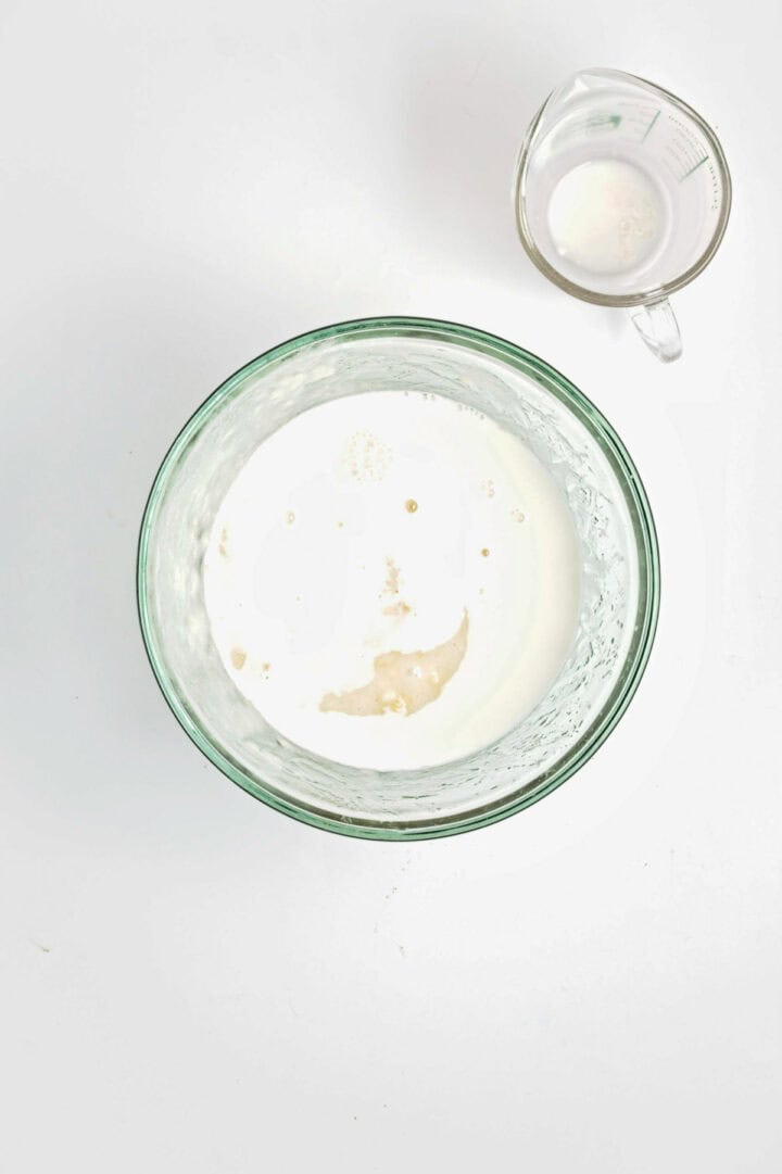 Glass bowl of milk and yeast mixture next to a clear measuring cup with liquid, on a white surface.