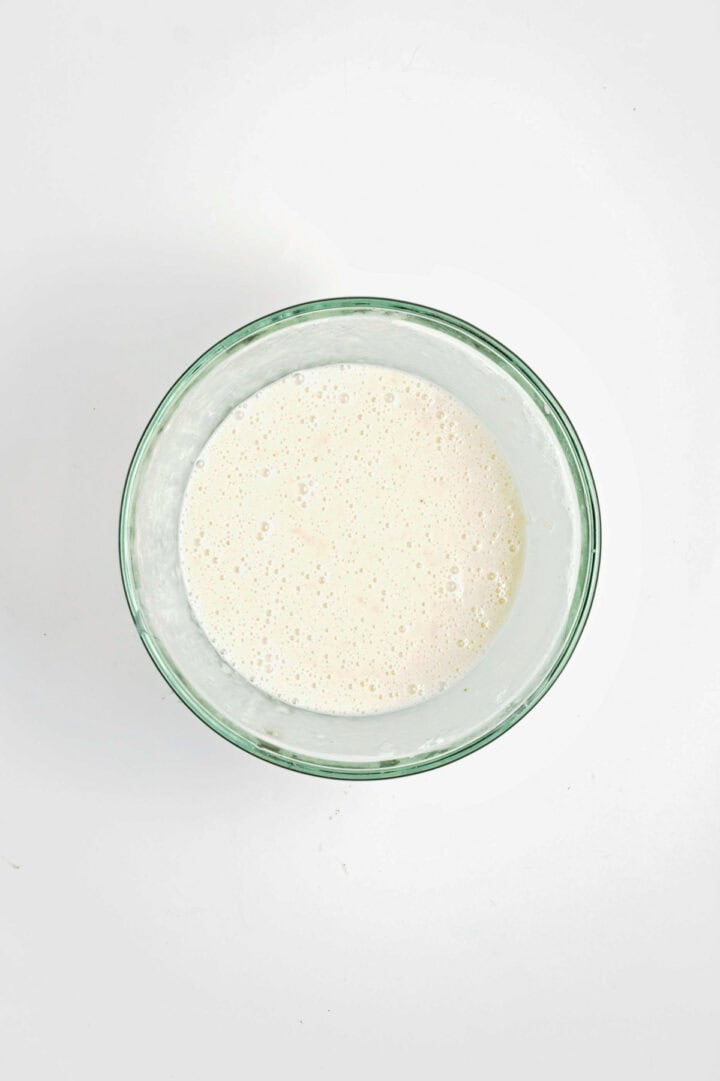 Glass bowl filled with a light, frothy liquid batter on a white surface, viewed from above.