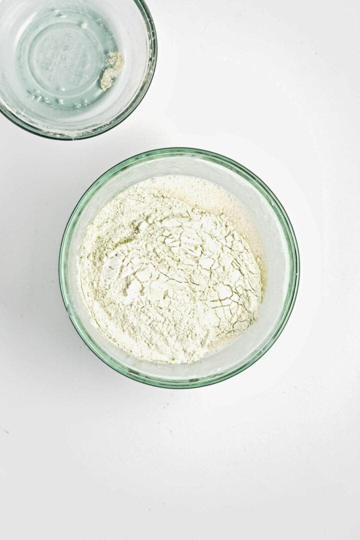 A glass bowl filled with flour sits on a white surface, with another empty bowl partially visible above.