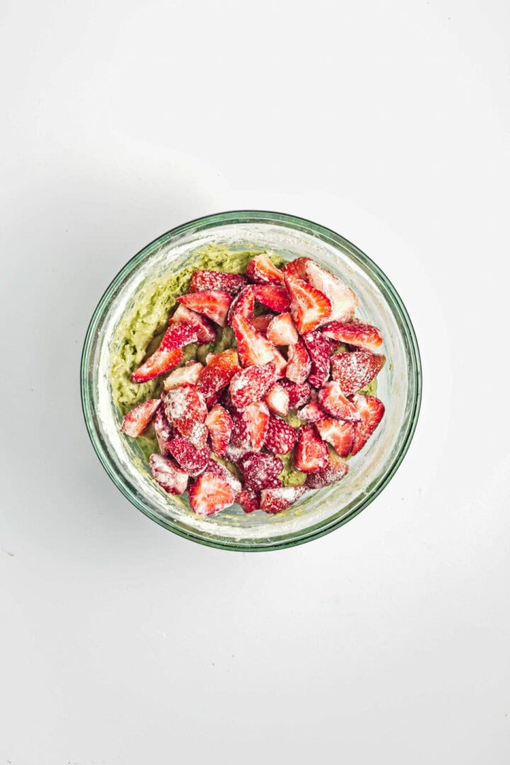 Sliced strawberries coated with sugar in a glass mixing bowl on a white surface.