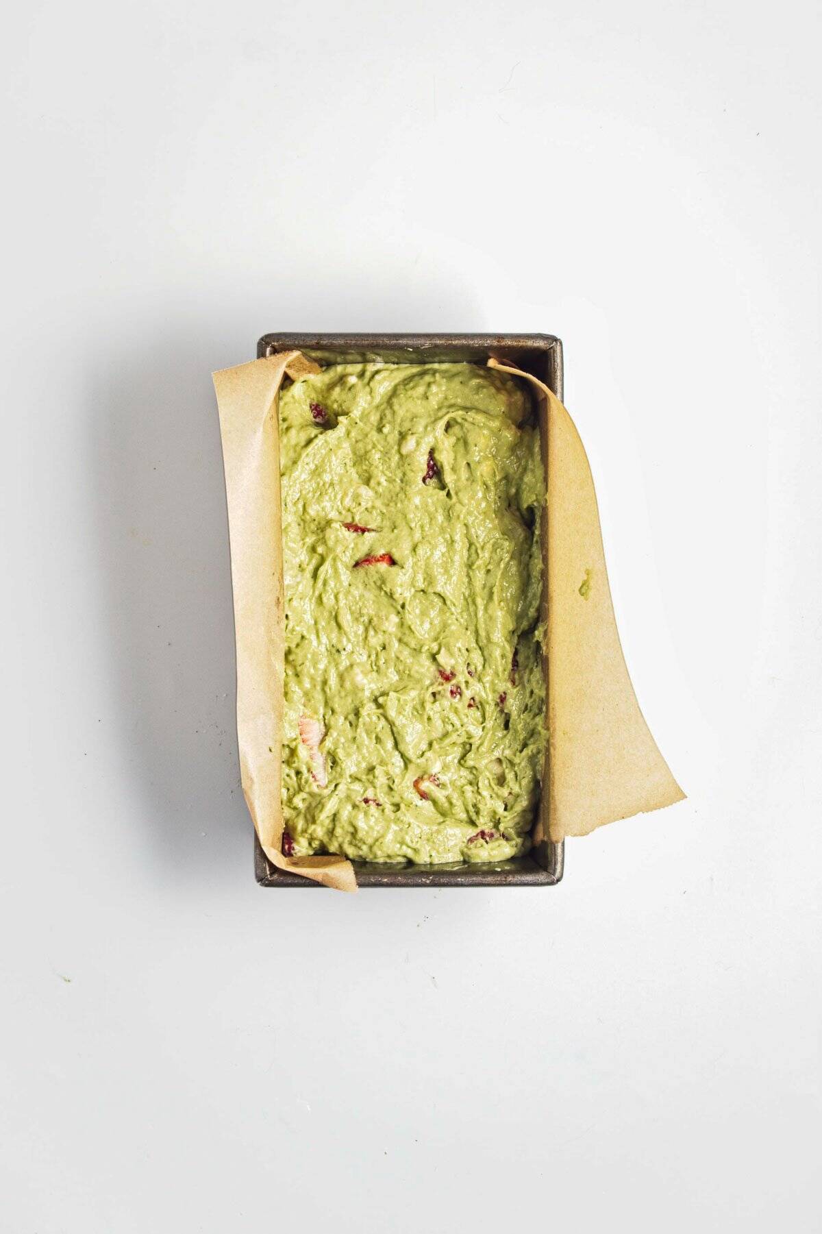 A loaf pan lined with parchment paper filled with green batter, viewed from above on a white surface.