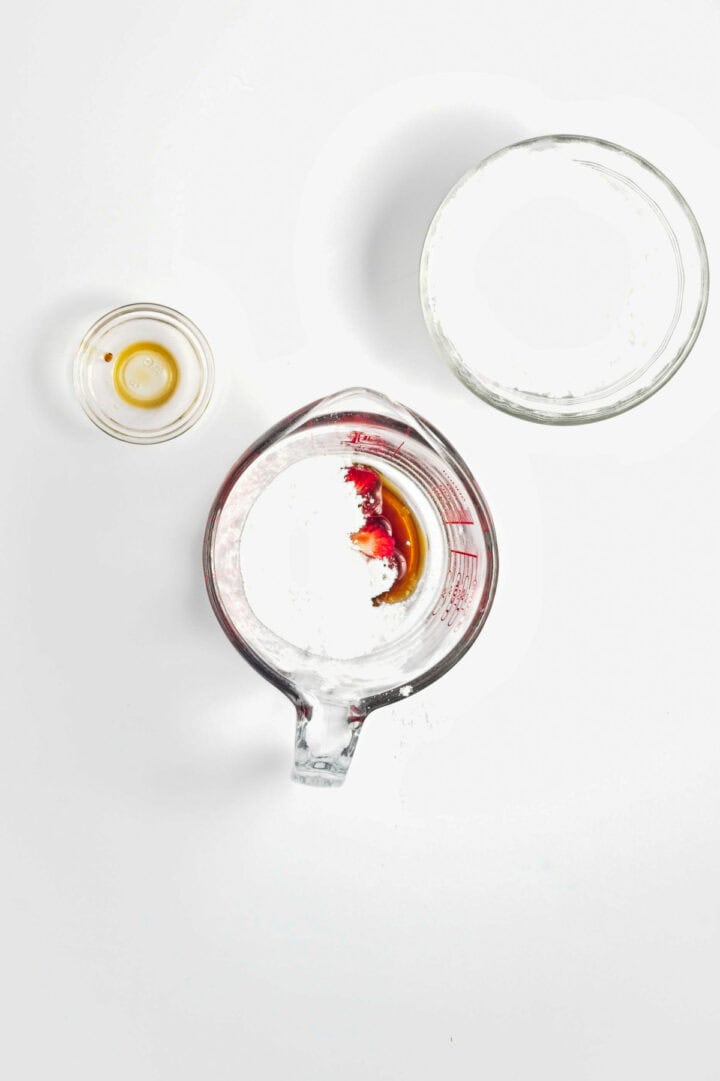 Glass bowls with liquid ingredients and a measuring cup with sugar, vanilla, and red food coloring on white background.