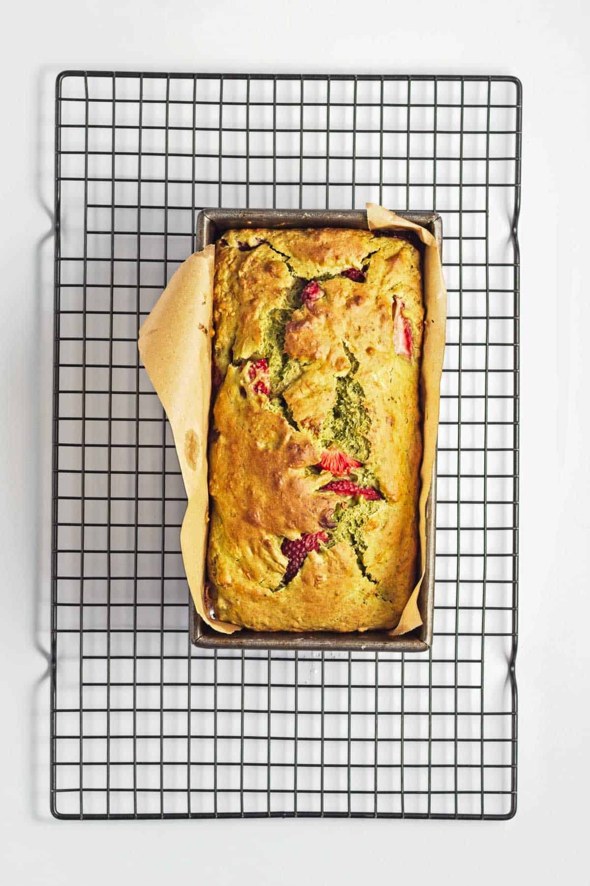 A loaf of bread with fruit pieces in a parchment-lined pan on a wire cooling rack.