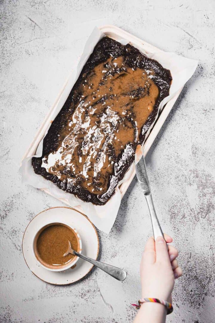 A hand spreads brown sauce over brownies in a parchment-lined pan, with more sauce in a dish nearby.