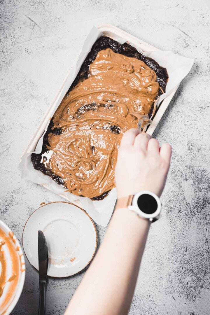 A hand spreads brown batter over dark brownie mix in a parchment-lined baking pan on a gray countertop.