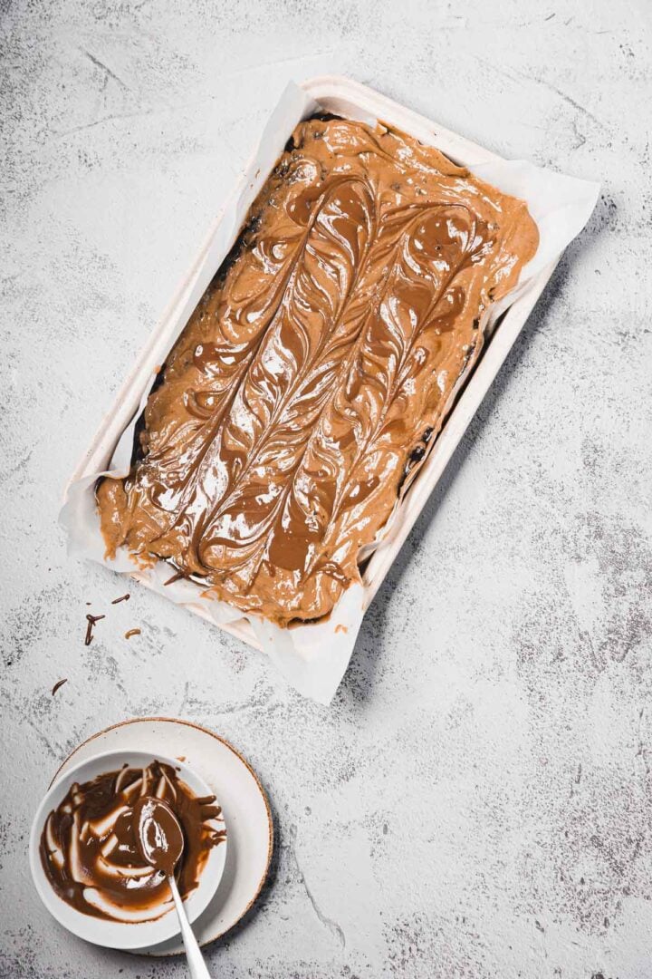 A pan of brownie batter swirled with chocolate, next to a small bowl of melted chocolate with a spoon.