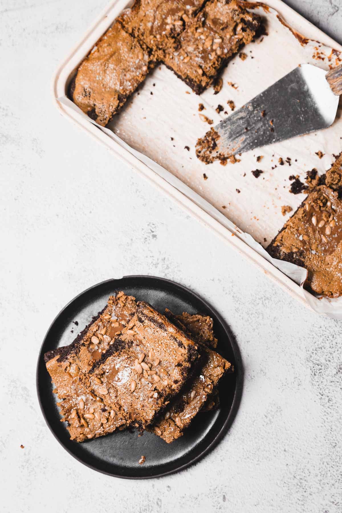 Two brownies on a black plate beside a baking dish with more brownies and a metal spatula.