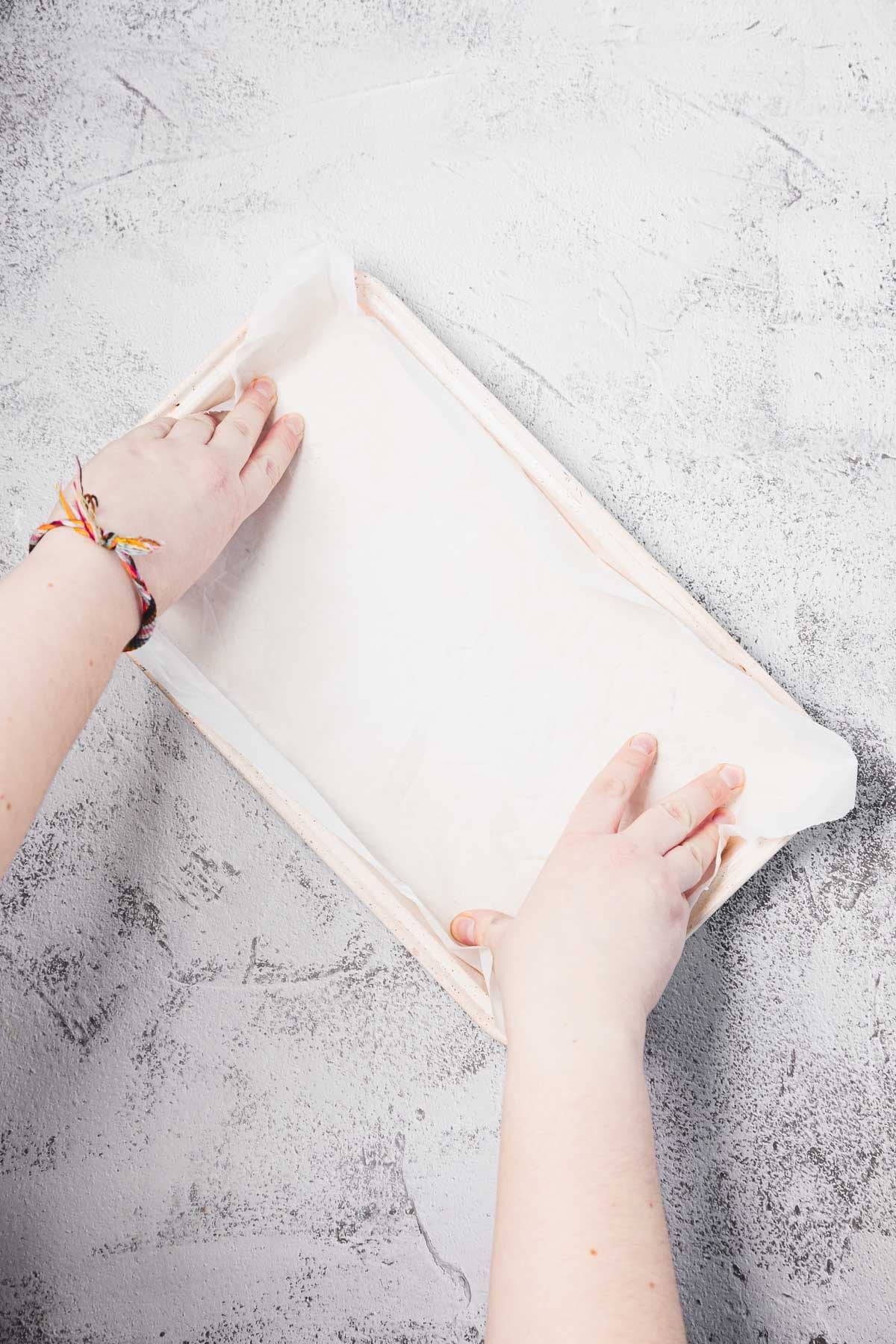 Hands lining a rectangular baking pan with parchment paper on a light textured surface.
