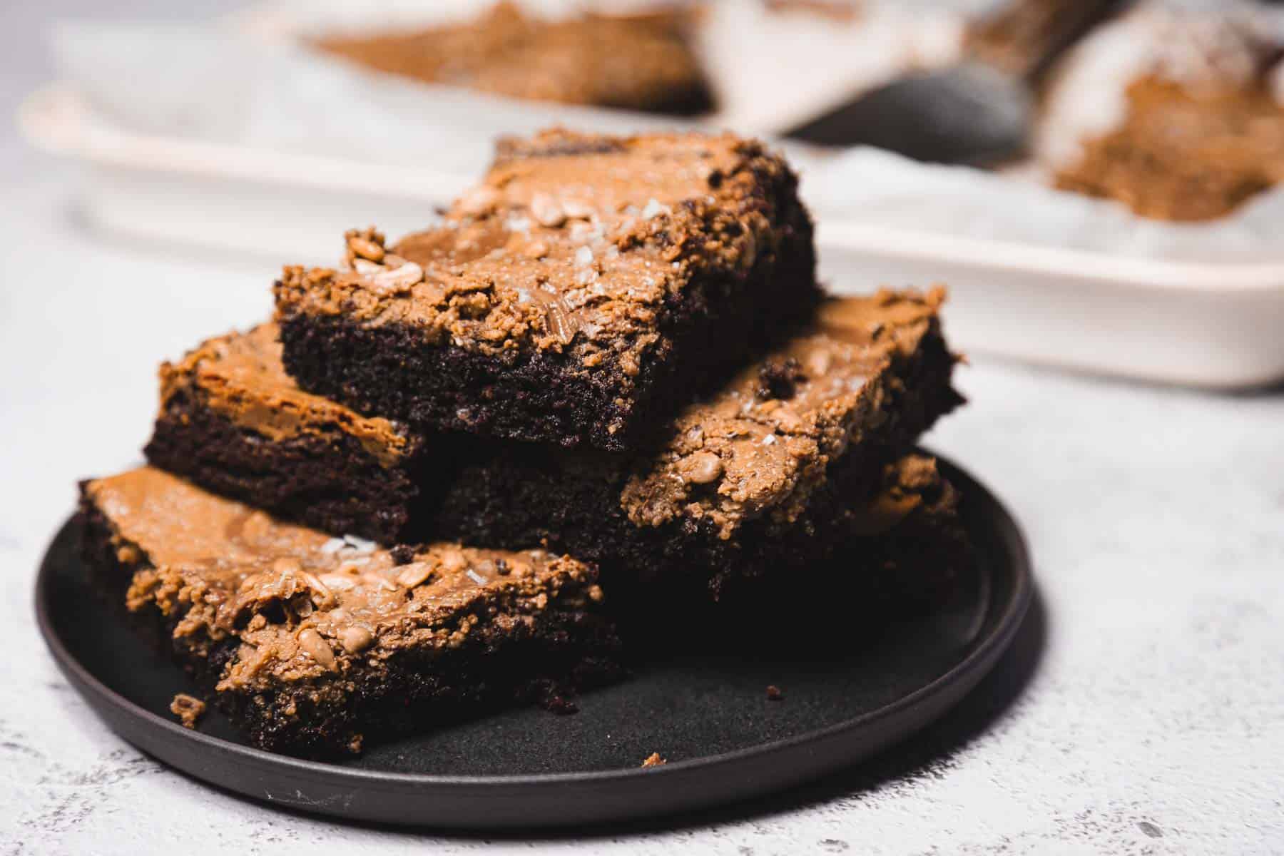 A stack of chocolate brownies sits on a dark plate, with more brownies in the background.
