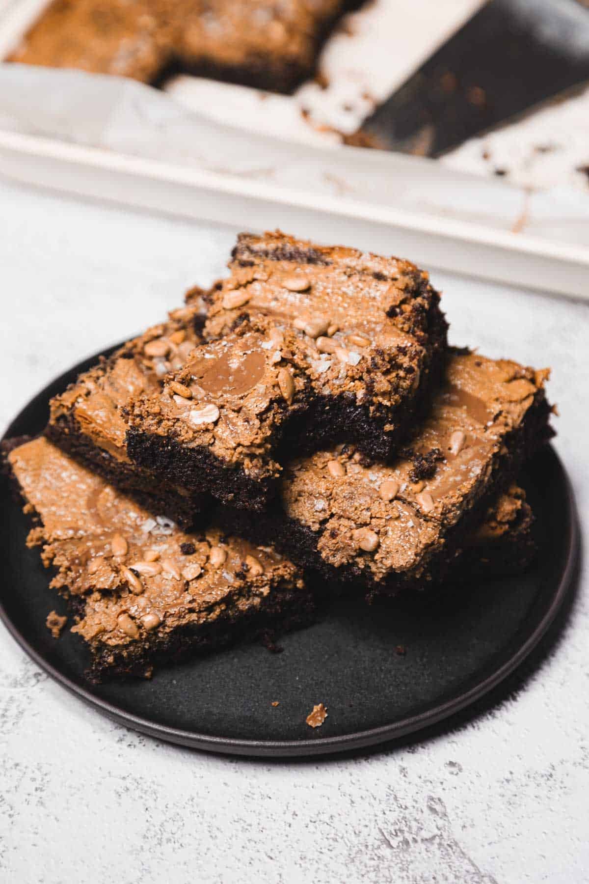 Three chocolate brownies stacked on a black plate, with more brownies visible in the background.