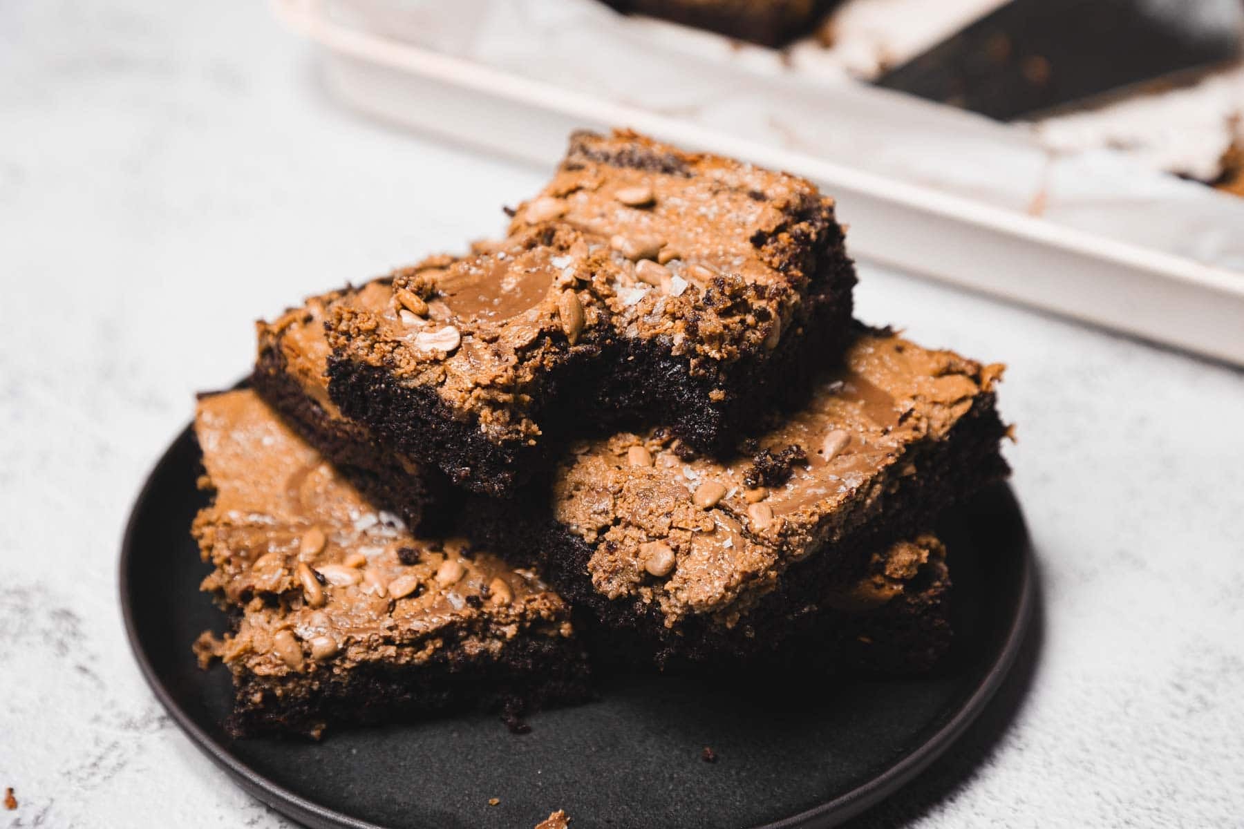 Three chocolate brownies stacked on a black plate, with more brownies in a tray in the background.