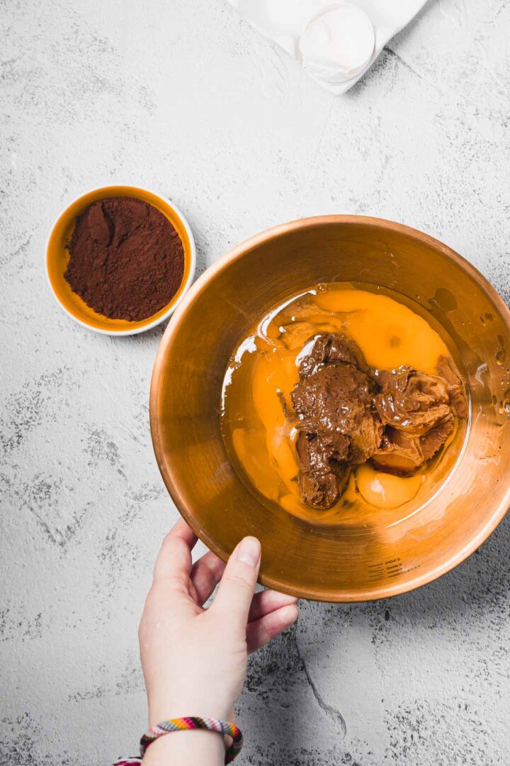 A hand holds a bowl with eggs and chocolate; a small bowl of cocoa powder is nearby on a gray surface.